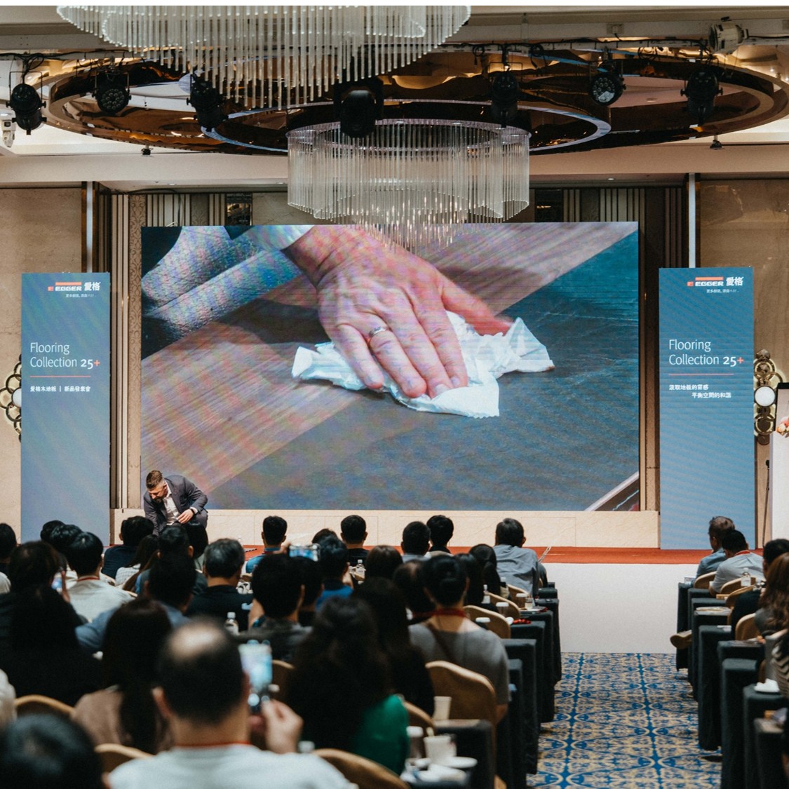 EGGER 講者於舞台進行木地板板材表面處理演示，投影畫面呈現產品耐污與易清潔特性，吸引參與者目光。