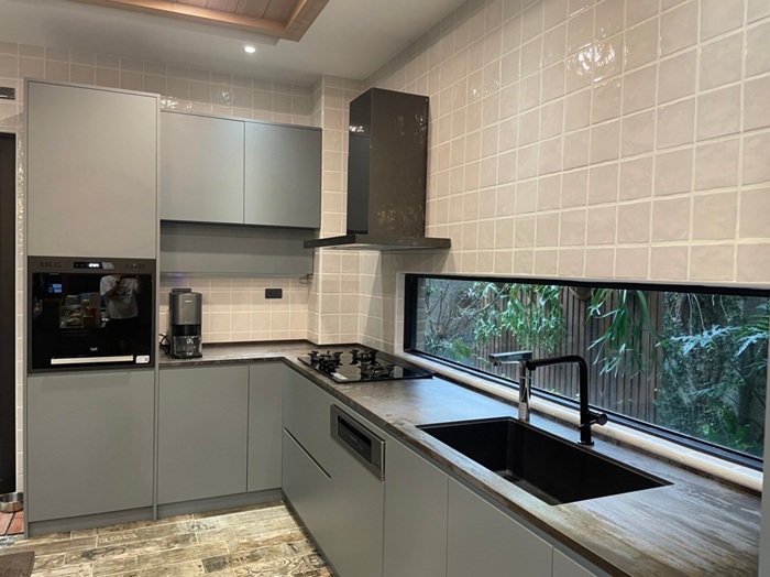 以 EGGER 灰色板材櫥櫃搭配木紋檯面與長形橫向窗，帶來自然採光與視覺延伸效果。廚房配備內嵌式烤箱、咖啡機及高品質龍頭，兼具美觀與功能性。